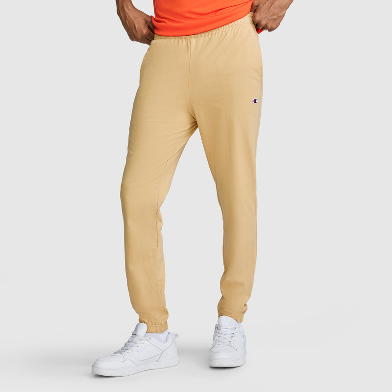 A person stands with hands resting on hips against a plain light-colored background, wearing Champion Closed Bottom Everyday Cotton Pants in beige crafted from 100% cotton, paired with a bright orange shirt and white sneakers.