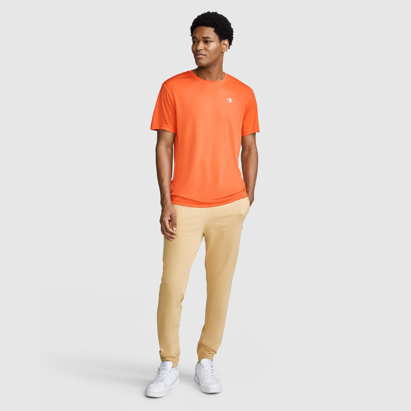 A man stands wearing an orange short-sleeve t-shirt, Champion Closed Bottom Everyday Cotton Pant in light beige, and white sneakers against a plain light gray background.