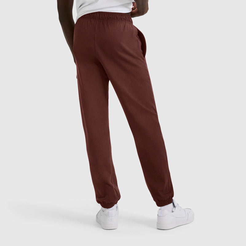 A person wearing Champion Closed Bottom Everyday Cotton Pant in maroon, paired with a white shirt and white sneakers, stands against a plain light gray background, facing away from the camera.