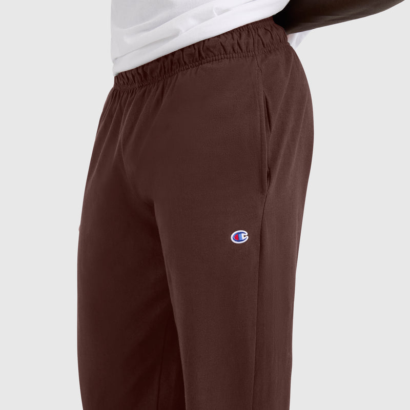 A person wearing brown Champion Closed Bottom Everyday Cotton Pants with an elastic waistband and side pockets. These comfortable pants have the Champion logo embroidered on the left thigh. The shirt is white, and the background is light gray.