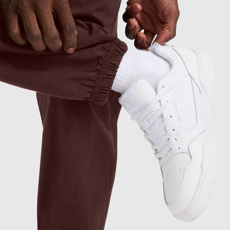 A person wearing Champion Closed Bottom Everyday Cotton Pant in brown, along with white socks and white sneakers, is adjusting the hem of their comfortable pants, focusing on the shoe and ankle area.