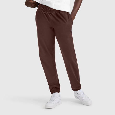 A person wearing Champion Closed Bottom Everyday Cotton Pants in dark brown, paired with a white t-shirt and white sneakers, stands with one hand in their pocket against a plain light gray background.