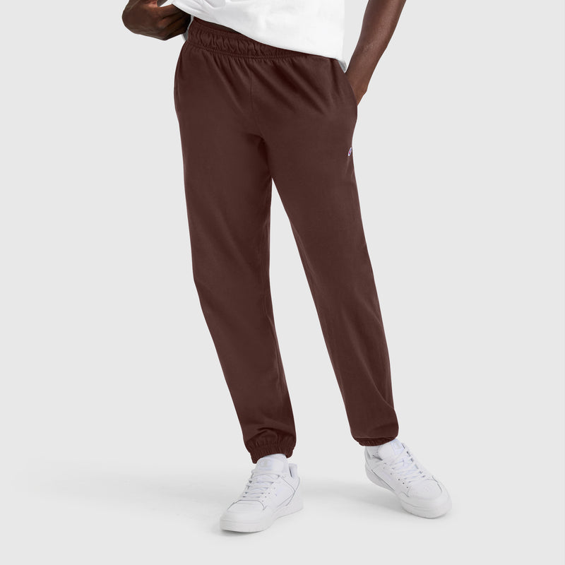 A person wearing Champion Closed Bottom Everyday Cotton Pants in dark brown, paired with a white t-shirt and white sneakers, stands with one hand in their pocket against a plain light gray background.