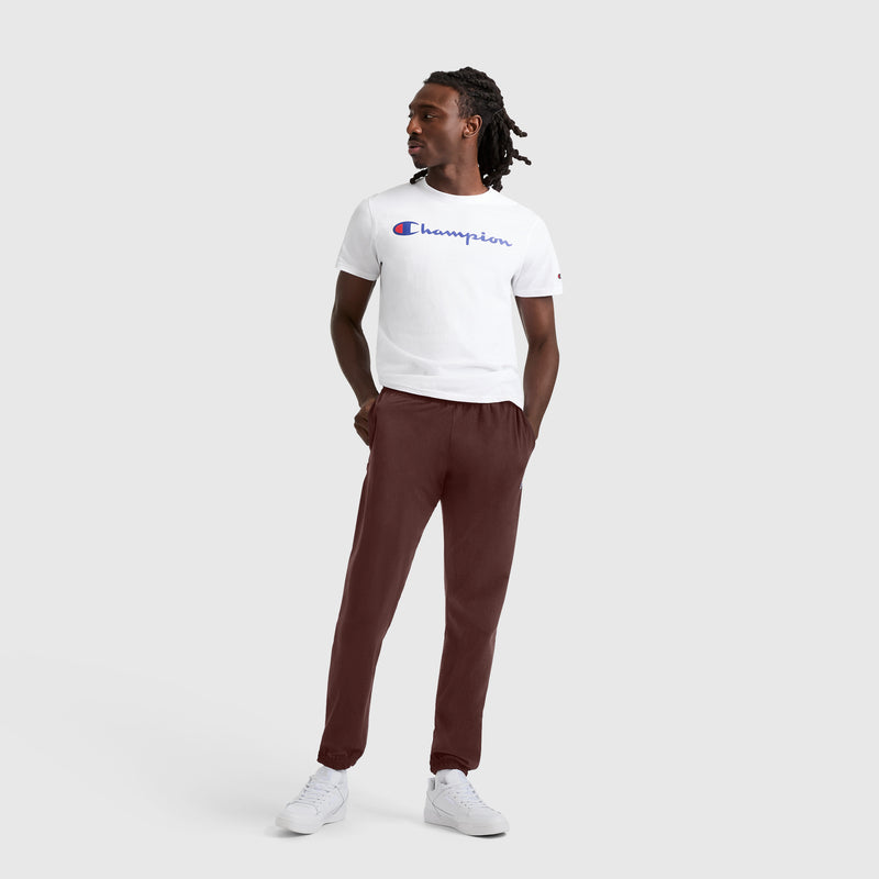 A person stands facing left against a plain light gray background, wearing a white Champion logo t-shirt, Champion Closed Bottom Everyday Cotton Pant in maroon, and white sneakers. Their hands are in their pockets, and they have shoulder-length braided hair.
