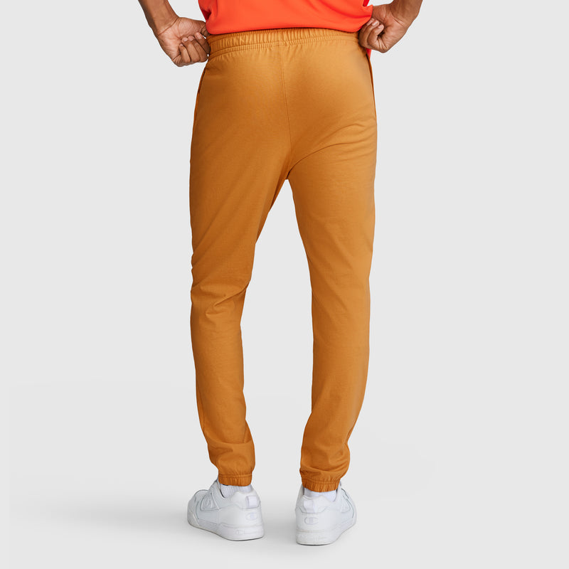 A person stands facing away, wearing Champion Closed Bottom Everyday Cotton Pants in light brown, paired with a bright orange shirt and white sneakers, against a plain light background.