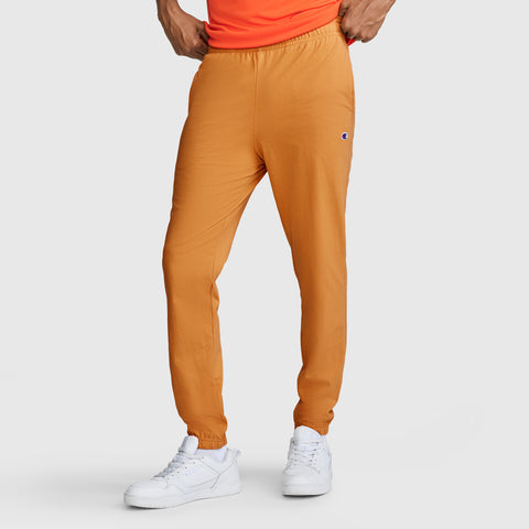 A person wearing Champion Closed Bottom Everyday Cotton Pants in orange stands against a plain light background, paired with a bright orange shirt and white sneakers. Only the lower half of the body is visible.