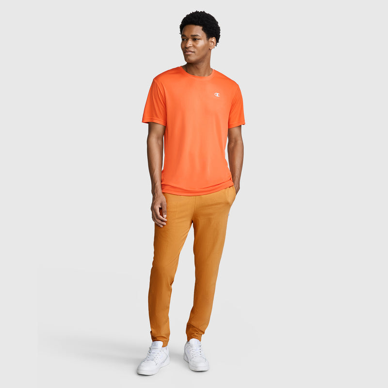 A man stands wearing an orange T-shirt, Champion Closed Bottom Everyday Cotton Pant in a tan shade, and white sneakers against a plain light gray background. He is looking to the side and smiling slightly.