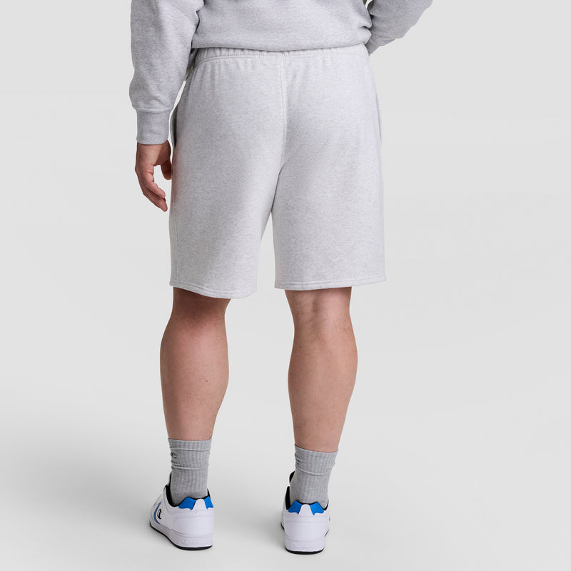 Person stands with their back to the camera in a light gray sweatshirt, Champion 10" Powerblend Fleece Short - Big & Tall, gray socks, and white sneakers with blue accents against a plain light background.