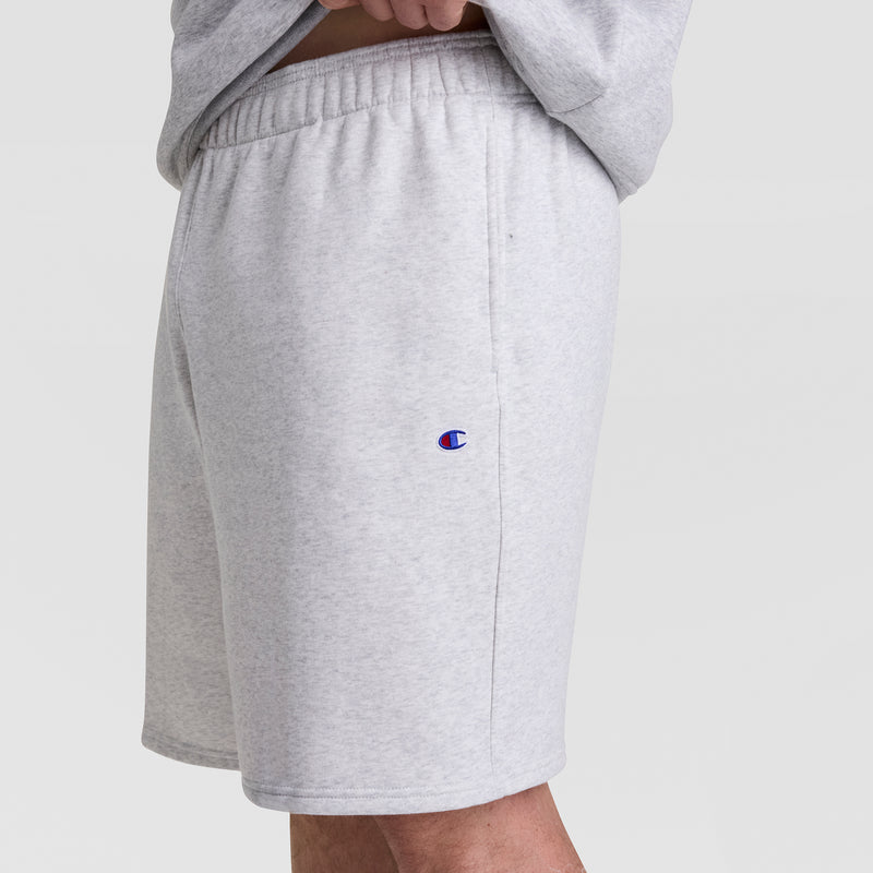 A person wears Champion 10" Powerblend Fleece Shorts - Big & Tall in light gray, featuring an elastic waistband and small logo on the left thigh. The side-view photo shows their lower torso and legs against a plain background.