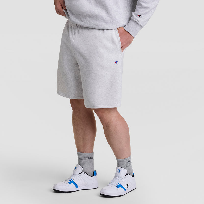 A person wearing Champion’s 10" Powerblend Fleece Short - Big & Tall in light gray, a matching sweatshirt, gray socks, and white sneakers with blue accents stands with one hand in their pocket against a plain white background.
