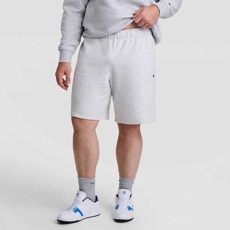 A person wearing Champion’s 10" Powerblend Fleece Short - Big & Tall in light gray, a matching sweatshirt, gray socks, and white sneakers with blue accents stands against a plain white background. Their face is not visible.