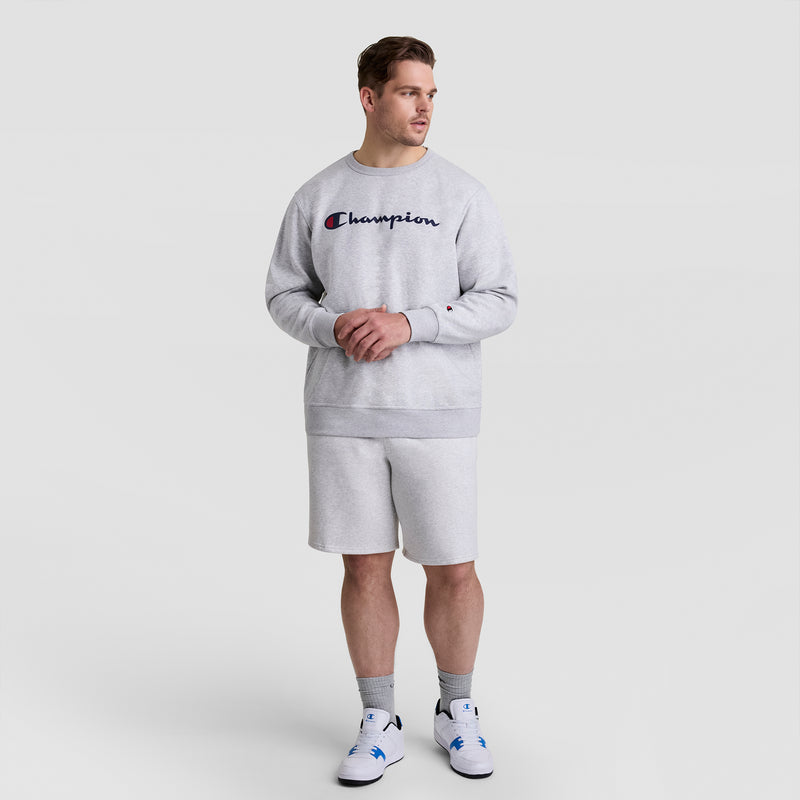 A man in a light gray Champion sweatshirt and 10" Powerblend Fleece Short - Big & Tall stands clasping his hands and looking sideways. He has short hair, gray socks, and white sneakers against a plain light gray background.