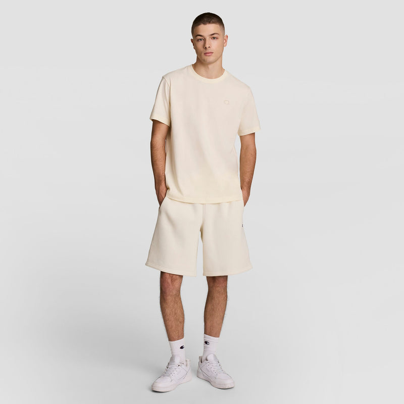 A young man wearing an off-white t-shirt, matching shorts, white socks, and white sneakers stands against a plain light gray background with his hands in his pockets.