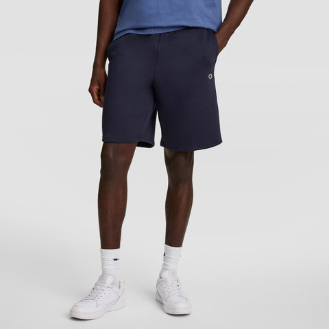 A person wearing a blue t-shirt, Champion 10" Powerblend Fleece Short, white socks, and white sneakers stands with one hand in their pocket against a plain light background.