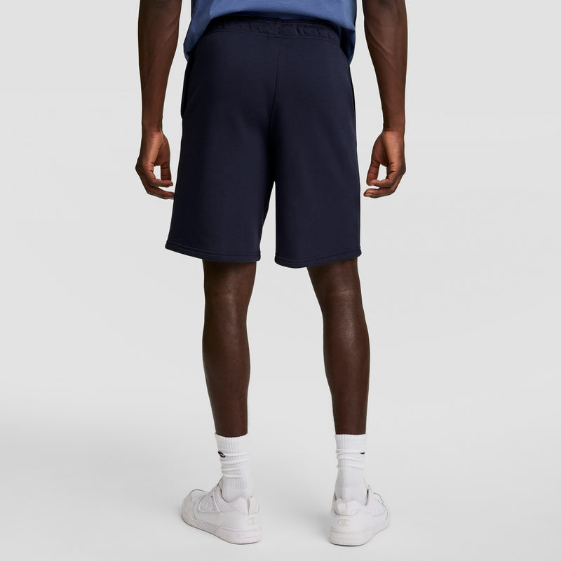 A person stands facing away in a blue shirt, Champion 10" Powerblend Fleece Shorts in navy, white crew socks, and white sneakers against a plain light background.