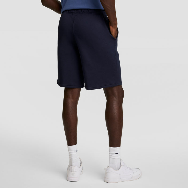 A person wearing Champion 10" Powerblend Fleece Shorts in navy, paired with white socks and sneakers, stands against a plain light gray background, shown from the waist down and facing away from the camera.