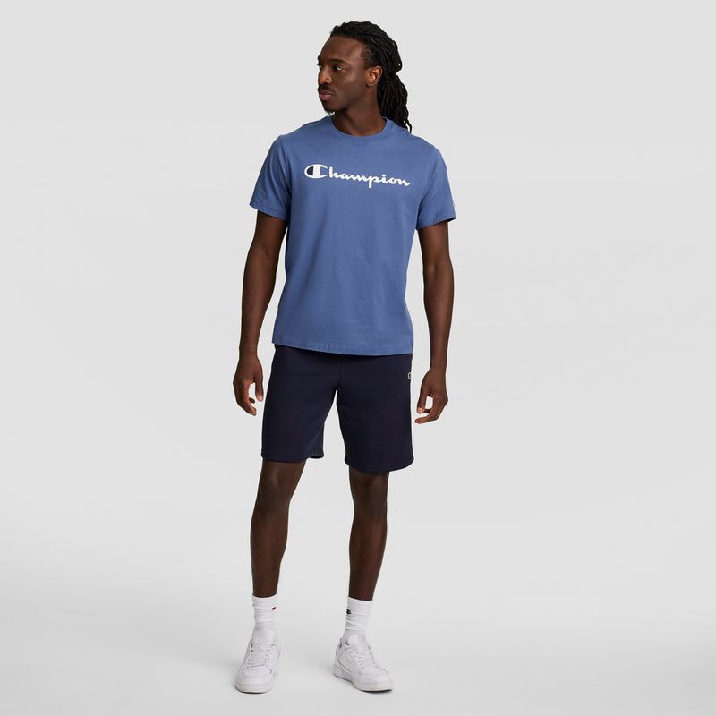 A man stands angled to the side, wearing a blue Champion t-shirt and Champion 10" Powerblend Fleece Short, paired with white socks and sneakers, set against a plain light gray background.