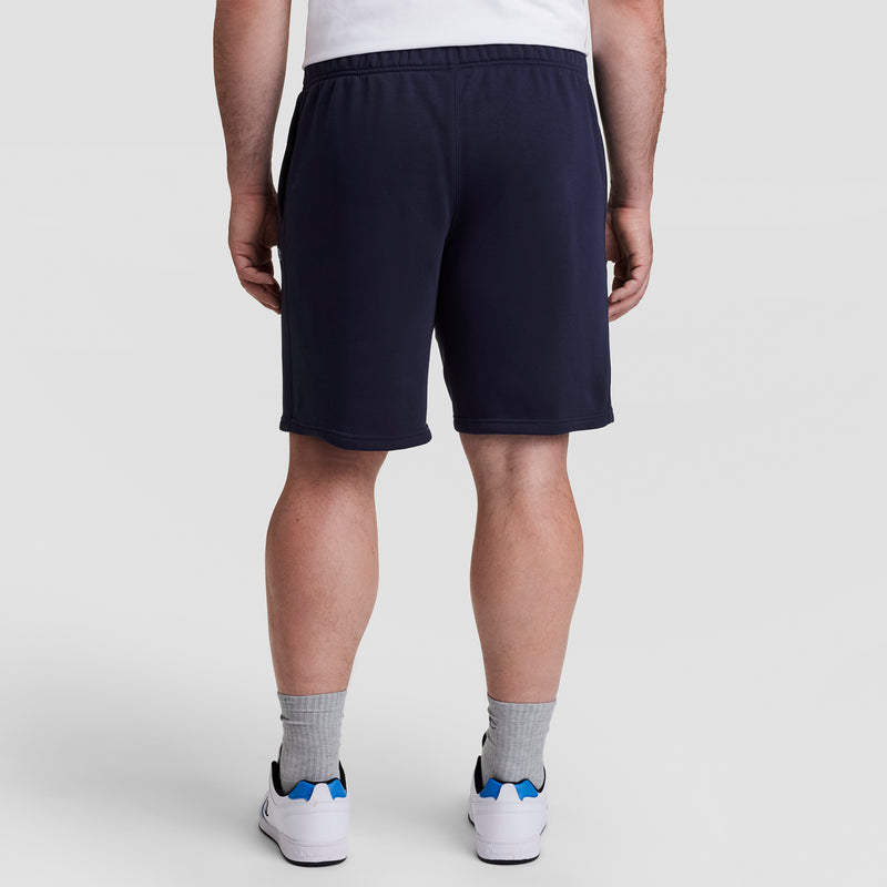 A person wearing a white shirt, Champion 10" Powerblend Fleece Short - Big & Tall in navy, gray socks, and white sneakers with blue accents stands facing away from the camera against a plain light background.