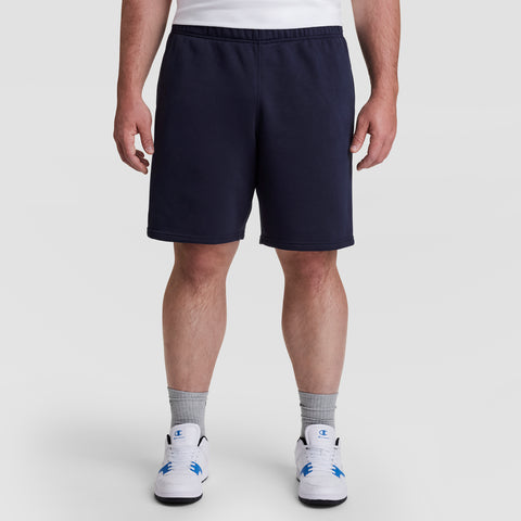 A person wearing a white T-shirt, Champion 10" Powerblend Fleece Short - Big & Tall in navy blue, gray crew socks, and white athletic shoes with blue accents stands against a plain light background.