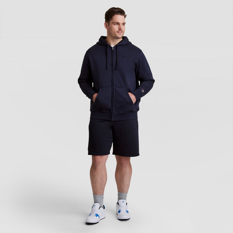 A man stands against a plain background wearing a navy zip-up hoodie, Champion 10" Powerblend Fleece Short - Big & Tall, gray socks, and white sneakers with blue accents. His hands are in his pockets as he looks slightly to the side.