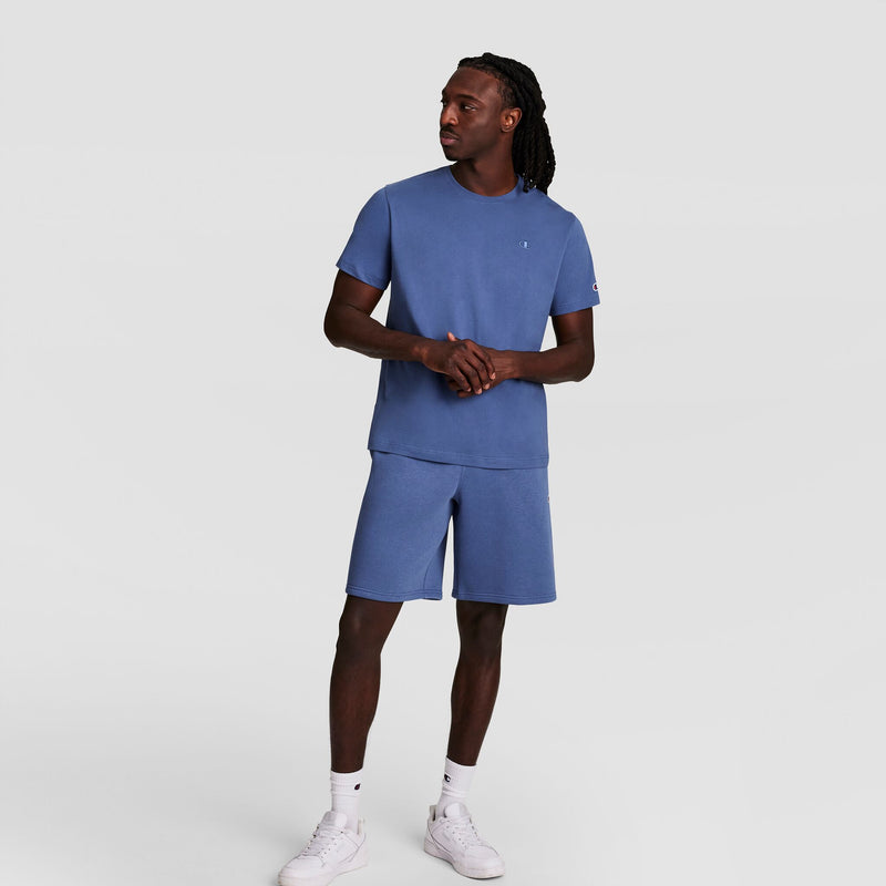 A man stands against a plain background, wearing a matching blue t-shirt, Champion 10" Powerblend Fleece Shorts, and white sneakers. He faces slightly to the side with his hands clasped together.