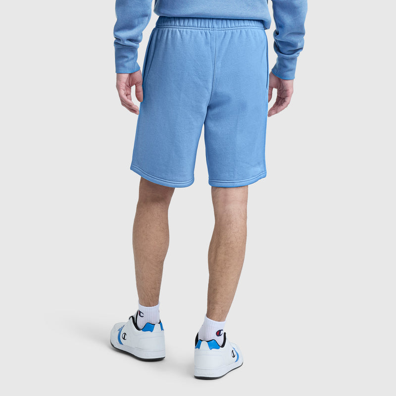 A person is shown from the back standing on a white background, wearing Champion’s 10″ Powerblend Fleece Shorts in light blue, paired with a matching sweatshirt, white socks featuring the blue and red Champion logo, and white sneakers.