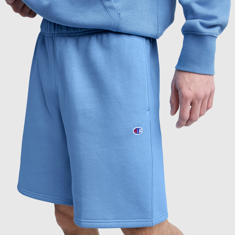 Wearing the Champion 10″ Powerblend Fleece Short in light blue, featuring the iconic Champion logo, and a matching Powerblend fleece sweatshirt, a person stands against a plain light background.