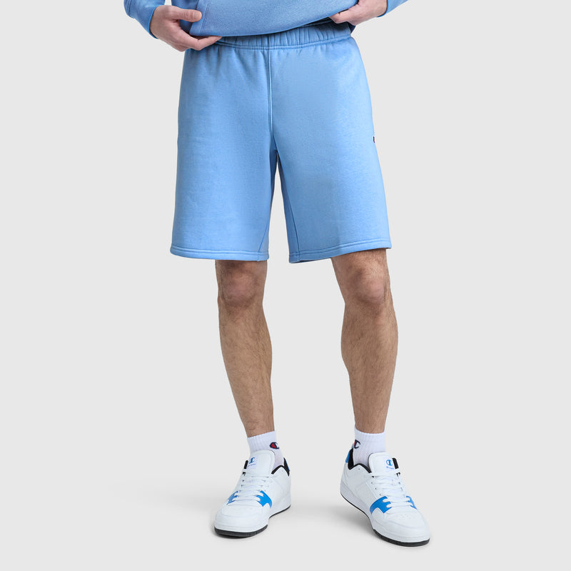 A person is shown from the waist down wearing Champion’s 10″ Powerblend Fleece Shorts in light blue, paired with white sneakers featuring blue accents, white socks, and a matching light blue sweatshirt. The background is plain and light-colored.