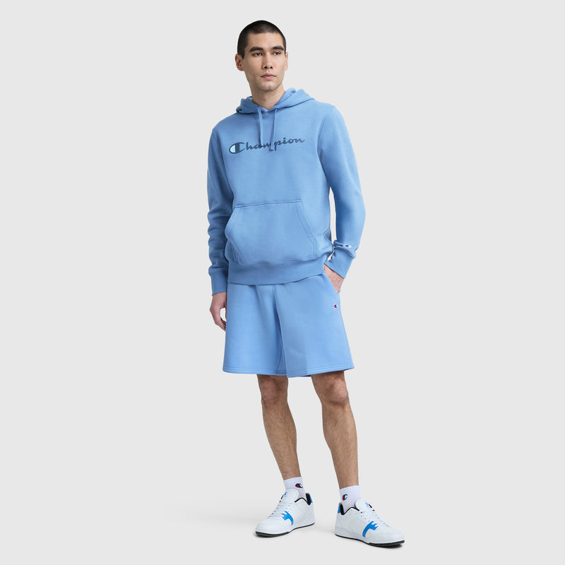 A person stands against a plain light background wearing a light blue Champion hoodie, Champion 10″ Powerblend Fleece Shorts, white socks, and white sneakers with blue details.