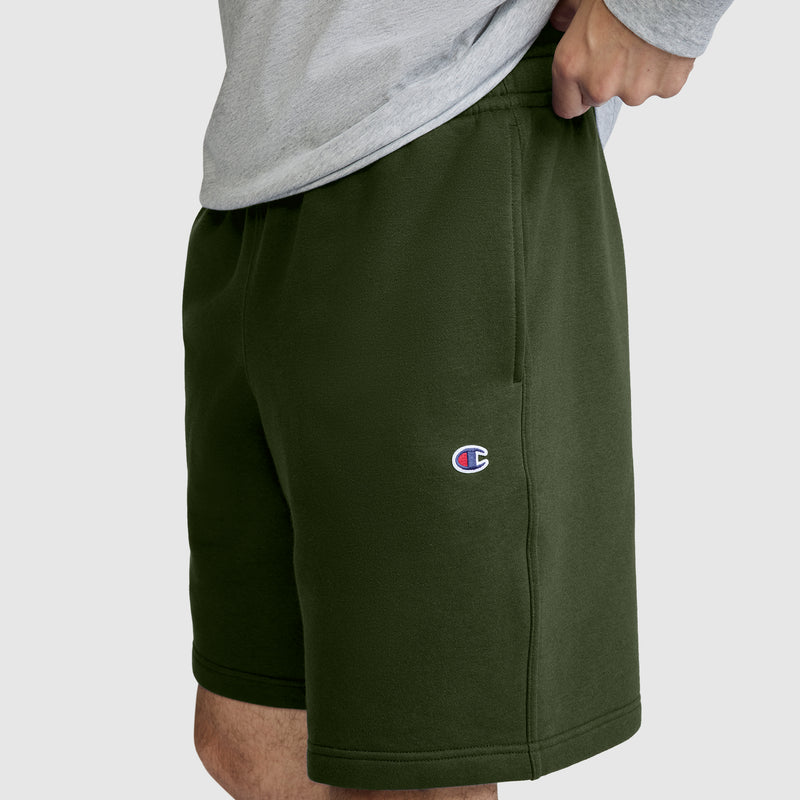 A person wearing olive green Champion 10" Powerblend Fleece Shorts and a gray shirt, with only the lower torso and upper legs visible. The Champion logo appears on the left leg of these men's shorts.