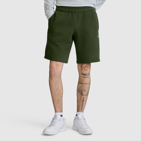 A person is wearing dark green Champion 10" Powerblend Fleece Shorts, a light gray long-sleeve shirt, and white sneakers. Only their lower body is visible, showing leg tattoos including a smiley face and a sword.