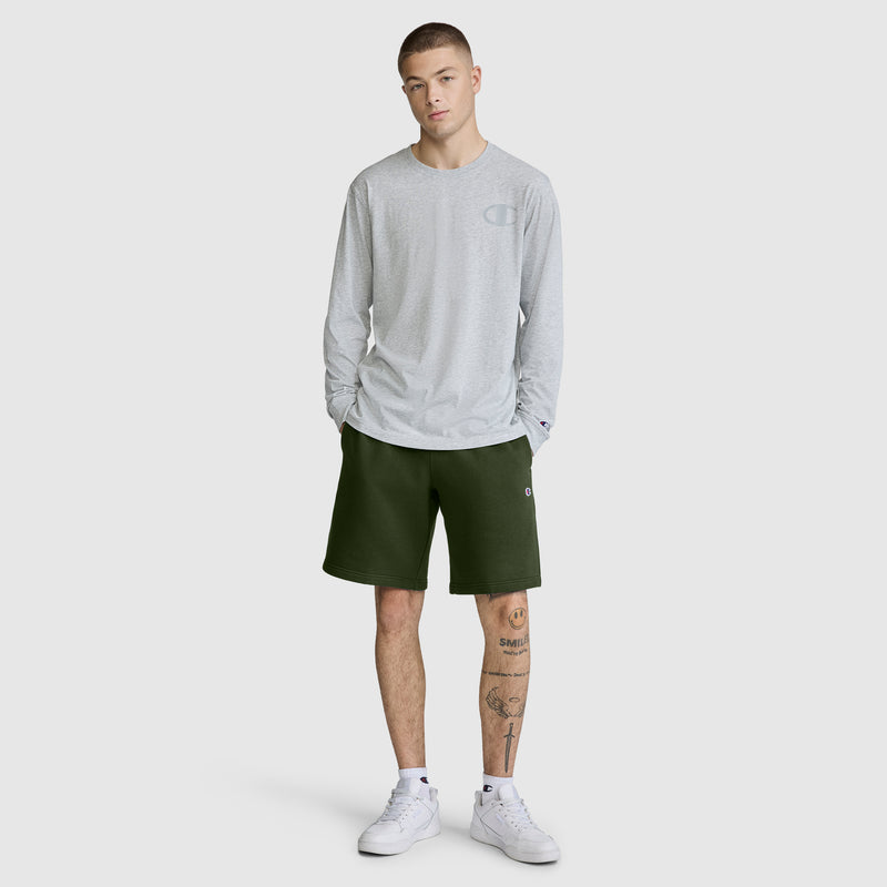 A person stands wearing a light gray long-sleeve shirt, the Champion 10" Powerblend Fleece Short in dark green, and white sneakers. They have short hair and a tattoo on their left leg. The background is plain and light-colored.