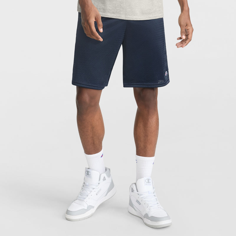 A person stands against a plain white background, showing only their lower body. They wear navy Champion 9" Classic Mesh Shorts with C Logo, a light gray shirt, white crew socks, and white athletic sneakers.