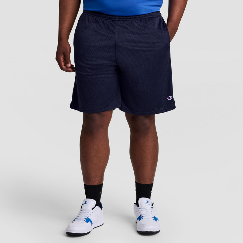 A person stands against a plain, light background, showing only their lower half while wearing Champion 9" Classic Mesh Shorts, C Logo - Big & Tall in navy, paired with a blue shirt, black socks, and white sneakers.