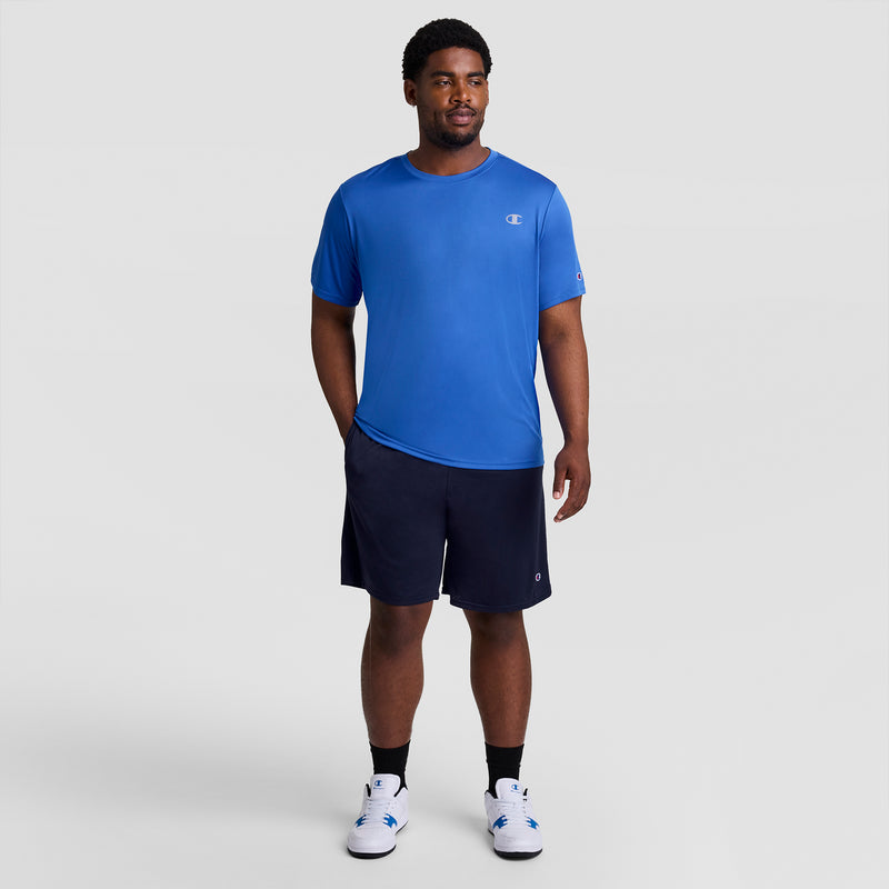 A man stands against a light gray background wearing a blue short-sleeve athletic shirt, white socks, and sneakers. He sports Champion 9" Classic Mesh Shorts, C Logo - Big & Tall in dark navy. His left hand is in his pocket as he looks slightly right.