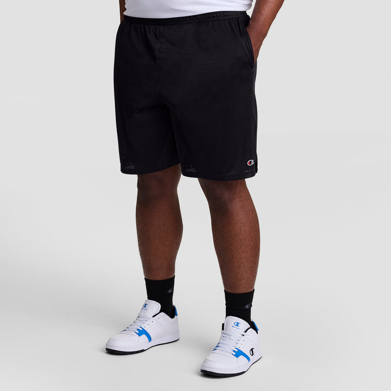 A person is shown from the waist down wearing Champion 9" Classic Mesh Shorts, C Logo - Big & Tall. They pair the black shorts with black socks and white sneakers with blue accents, standing against a plain light background.
