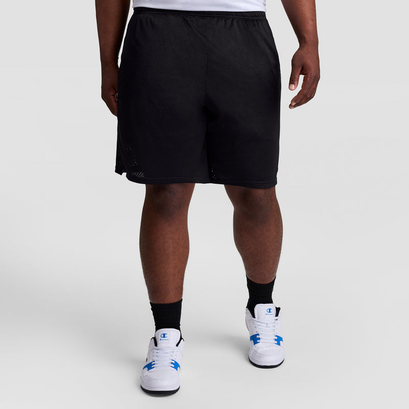 A person is seen from the waist down wearing Champion 9" Classic Mesh Shorts, C Logo - Big & Tall in black, along with black socks and white sneakers with blue accents, standing against a plain gray background.