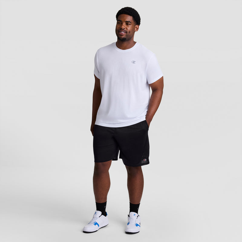 A man stands against a light gray background, wearing a white short-sleeve T-shirt, Champion 9" Classic Mesh Shorts with C Logo - Big & Tall in black, black socks, and white sneakers with blue accents. Hands in pockets, he smiles.