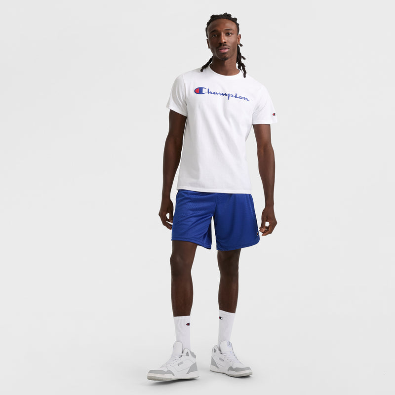 A man with long braided hair and a neutral expression stands against a white background, wearing a white Champion tee, Champion 9" Classic Mesh Shorts with C Logo in blue, white crew socks, and white sneakers. His arms are relaxed at his sides.