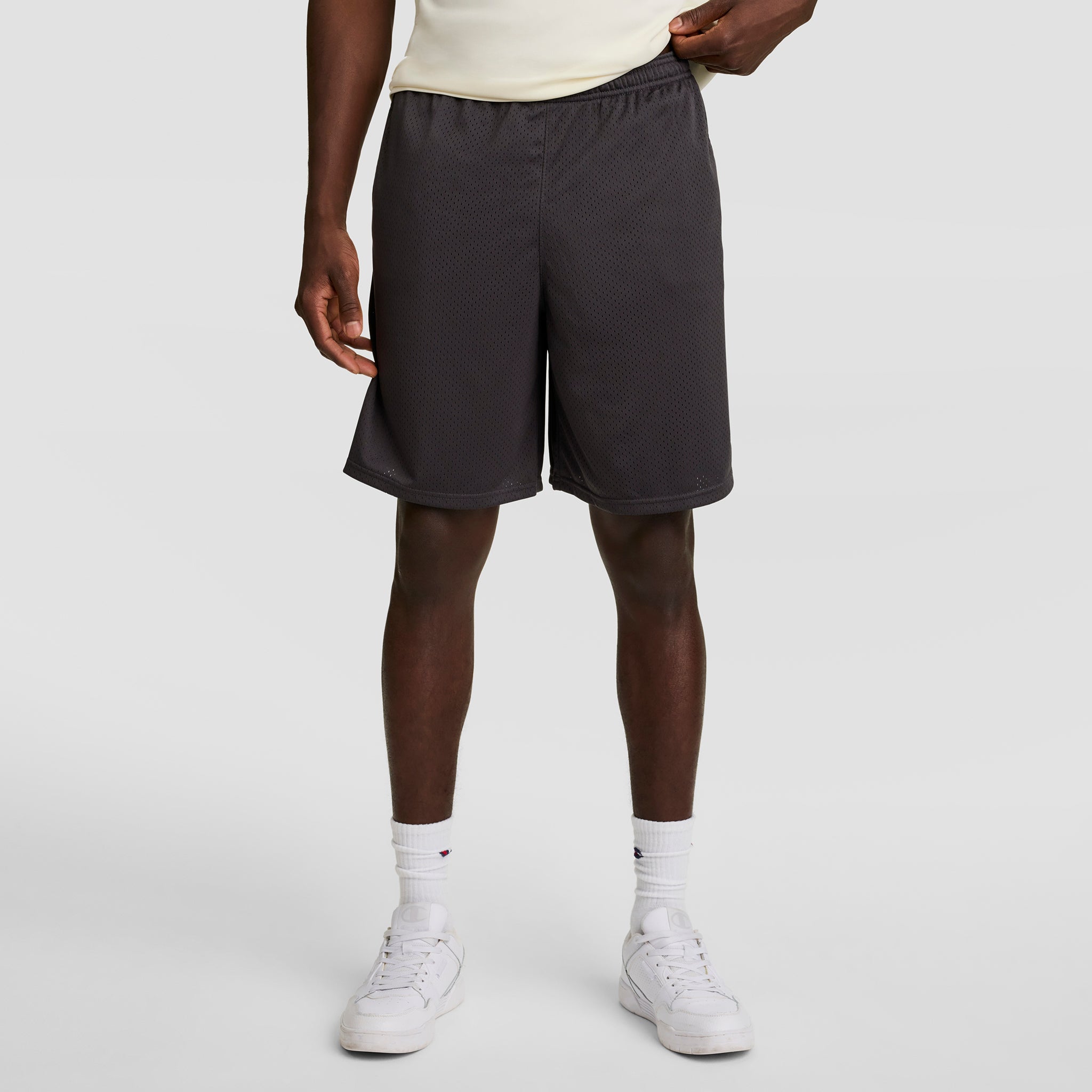 A person, visible from chest to feet, stands against a plain white background wearing a cream shirt, white crew socks, white sneakers, and Champion 9" Classic Mesh Shorts with C Logo in dark gray.