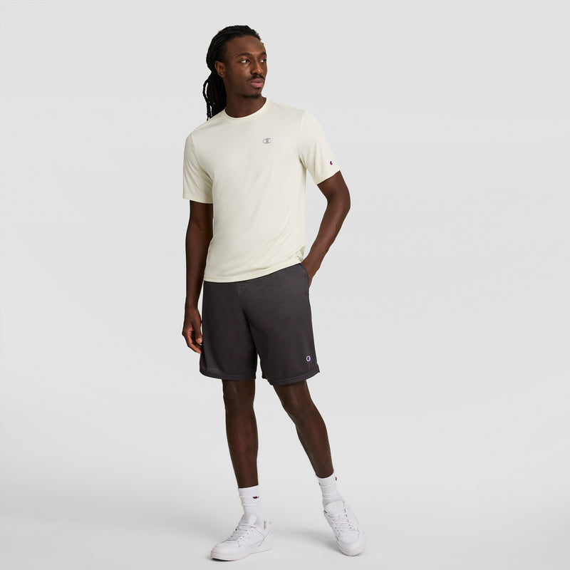 A man stands against a light gray background, wearing a light beige T-shirt, Champion 9" Classic Mesh Shorts with C Logo in dark gray, white socks, and white sneakers. He has medium-length twisted hair and looks to the side with one hand in his pocket.