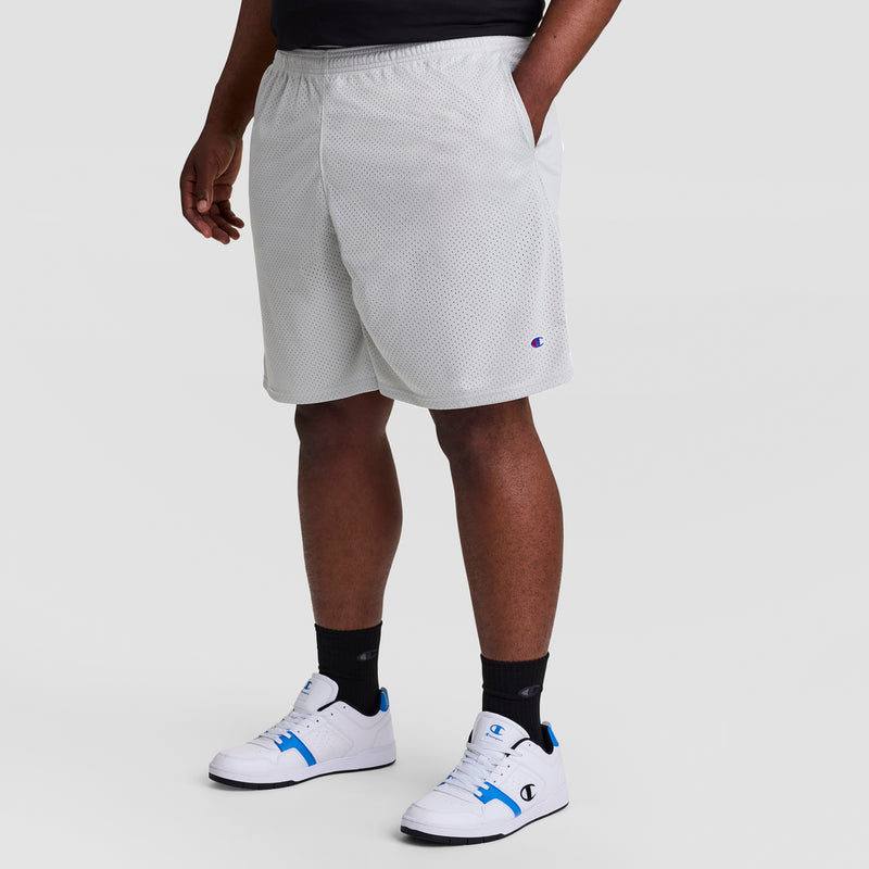 A person wears Champion 9" Classic Mesh Shorts, C Logo - Big & Tall, styled with a black shirt, black socks, and white sneakers with blue accents. They stand hands in pockets against a plain light background.