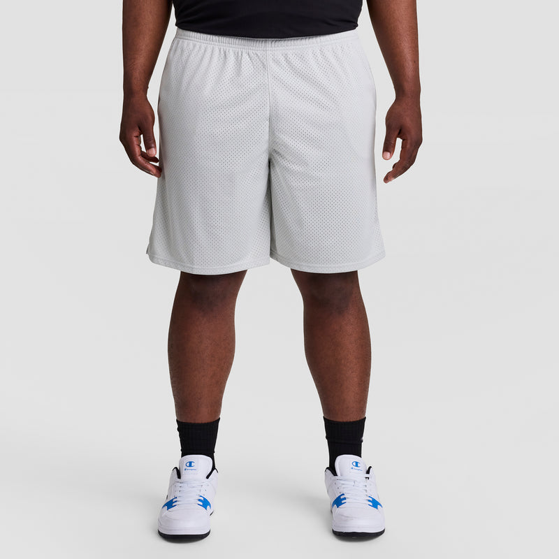 A person models the Champion 9" Classic Mesh Shorts, C Logo - Big & Tall in white, paired with black socks and white sneakers with blue accents, standing against a plain light gray background. The image is cropped waist down.