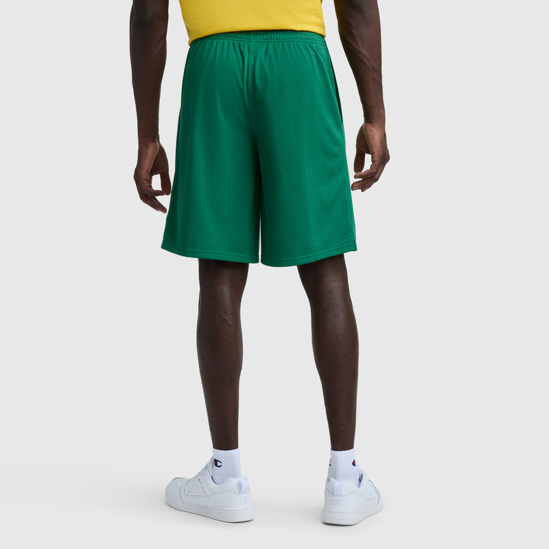 A person in a yellow shirt, white crew socks, and white sneakers faces away, displaying the back of green Champion 9" Classic Mesh Shorts, C Logo against a plain white background.