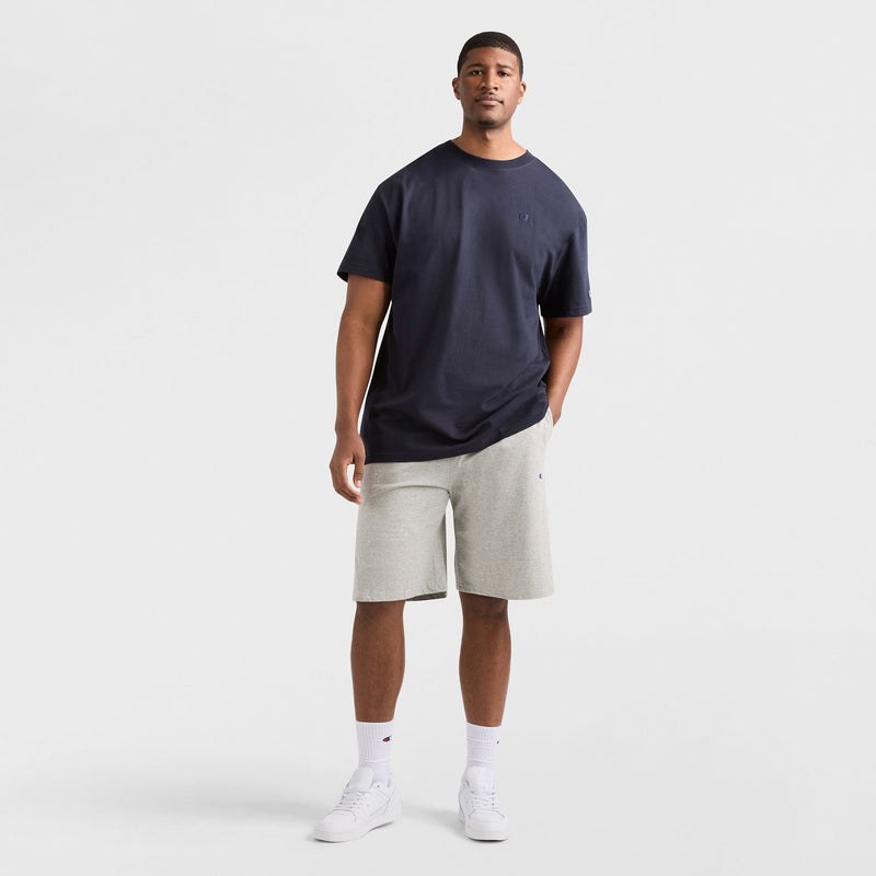 A man stands against a white background wearing a dark navy t-shirt, white socks, and white sneakers. He is also wearing the Champion 9" Lightweight Lounge Shorts with C Patch Logo - Big & Tall in light gray cotton jersey. One hand is in his pocket as he faces the camera with a neutral expression.