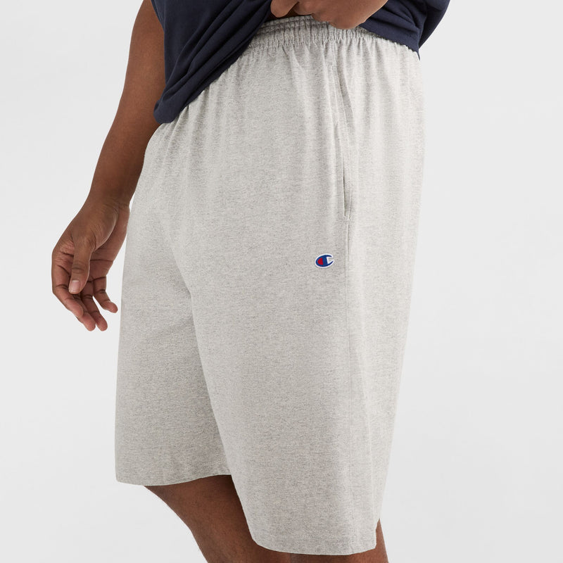A person is wearing light gray Champion 9" Lightweight Lounge Shorts with the C Patch Logo - Big & Tall, paired with a dark shirt. One hand rests at their side while the other lifts the shirt slightly, revealing the visible Champion logo on the lounge shorts.