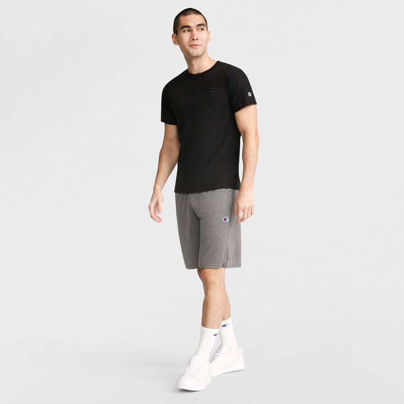 A man wearing Champion 9" Lightweight Lounge Shorts with the C Patch Logo in gray, paired with a black t-shirt, white socks, and white sneakers, stands against a plain light background. He is looking slightly to the side with one arm relaxed at his side and the other slightly bent.