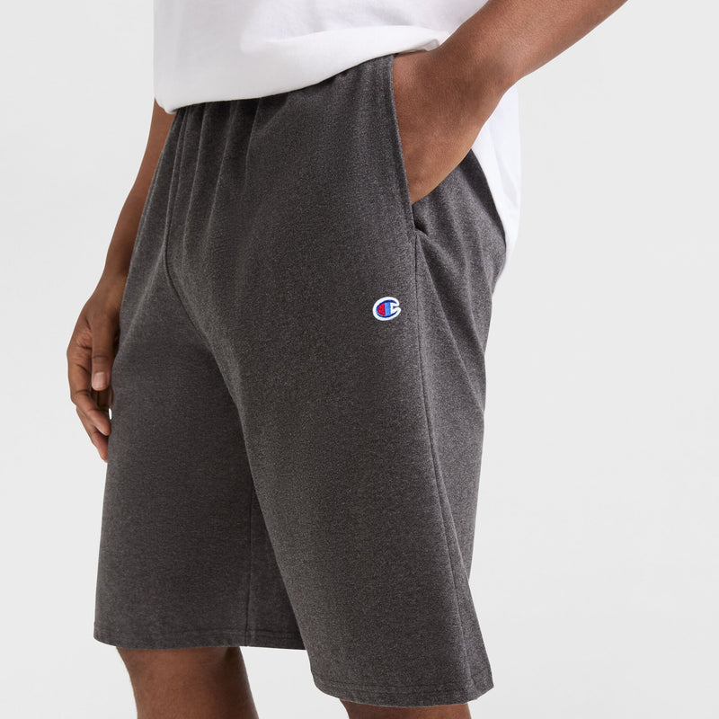 A person wearing a white shirt and Champion 9" Lightweight Lounge Shorts, C Patch Logo - Big & Tall in dark gray cotton jersey, with hands in pockets, shown from the waist down against a plain background.