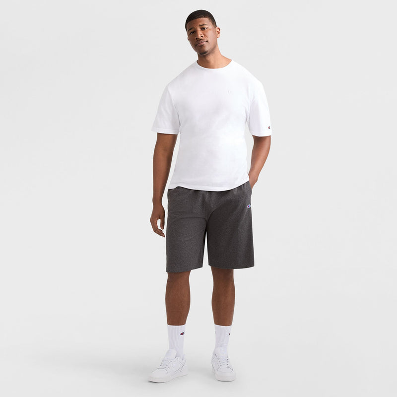 A man stands against a plain white background wearing a white T-shirt, Champion 9" Lightweight Lounge Shorts with C Patch Logo in dark gray (Big & Tall), white socks, and white sneakers. One hand is in his pocket and he is looking forward.