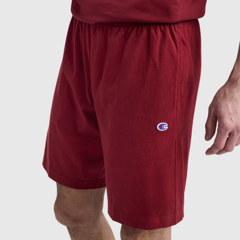 A person wears Champion 9" Lightweight Lounge Shorts, C Patch Logo in red with an elastic waistband and a small Champion logo on the left thigh. The shorts are perfect for lounging. Their right arm hangs by their side, paired with a matching red shirt.